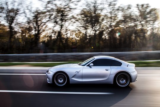 A silver metallic color coupe driving by the forest.