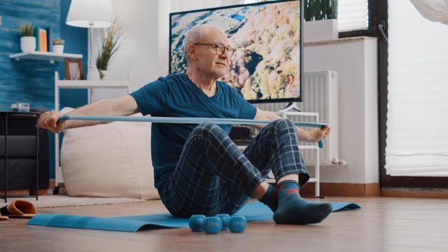 Old man using resistance band to stretch and pull, sitting on yoga mat. senior person training with elastic belt to do arms muscles exercise at home. elder adult doing physical activity