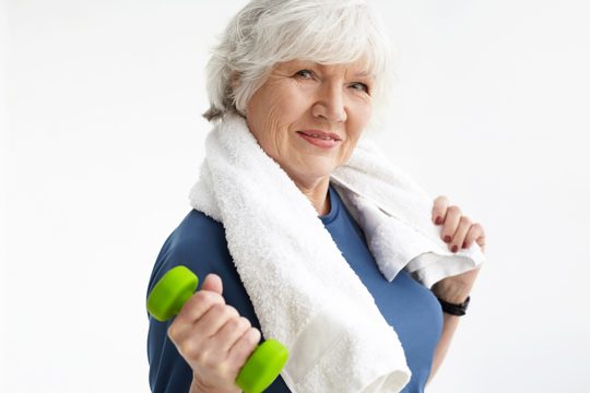 Strength, energy, wellness and healthy active lifestyle concept. stylish athletic senior female with fit body and gray hair wrking out in gym using dumbbell, wearing white towel around her neck