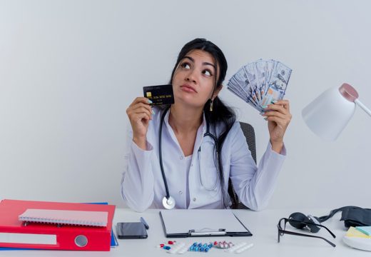 Thoughtful young female doctor wearing medical robe and stethoscope sitting at desk with medical tools holding money and credit card looking at side isolated