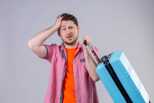 Young traveler man standing with suitcase looking at camera confused touching head for mistake over white background