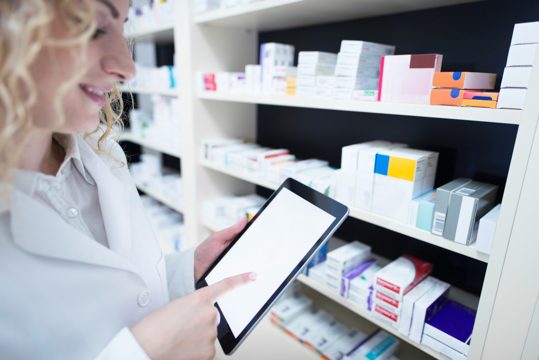Pharmacist holding tablet by the shelf full of medicine in drug store and checking medication availability and specification