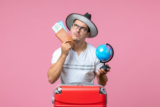 Front view of young man holding vacation tickets and little globe on pink wall