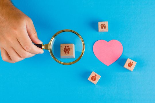 Family concept with paper cut heart on blue table flat lay. hand holding magnifying glass over wooden cube.