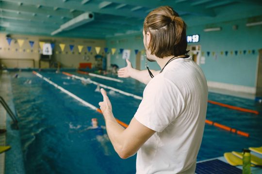Male swimming coach standing by the swimming pool. he is showing staight hand to correct the student. swimming technique, professional.