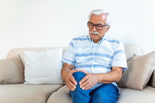 Elderly man sitting on a sofa at home and touching his painful knee people health care and problem concept unhappy senior man suffering from knee ache at home