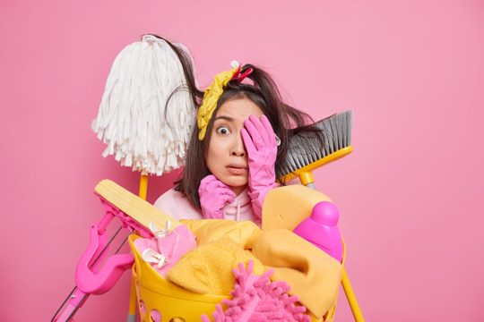 Scared asian woman keeps hand on face looks frightened at camera surrounded by cleaning tools afraids of starting to clean up very dirty room does laundry at home isolated on pink studio wall