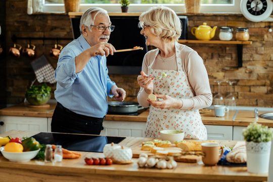 Happy mature man giving his wife to try out food he is cooking in the kitchen