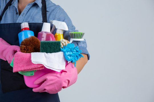 Young girl is holding cleaning product, gloves and rags in the basin on white wall