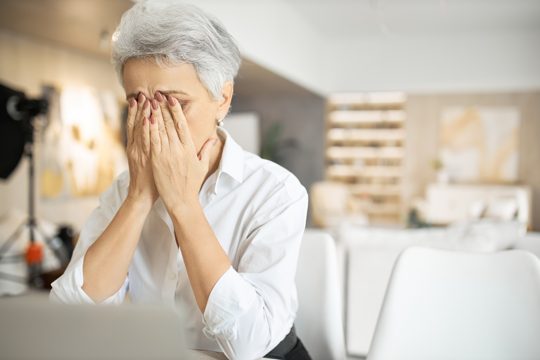 Senior sad woman with gray hair working on laptop, rubbing eyes or hiding tears, full of restless thoughts