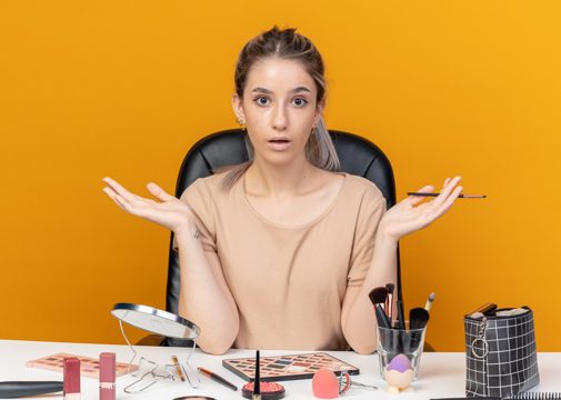 Surprised young beautiful girl sits at table with makeup tools holding makeup brush spreading hands isolated on orange background