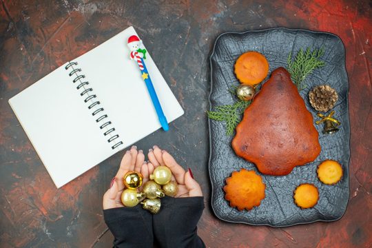 Top view cake and cupcakes on black plate xmas ball toys in female hands notebook and pen on dark red table