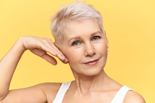 Portrait of attractive middle aged caucasian woman in tank top touching cheek, applying anti wrinkle moisturizing cream, looking at camera with smile.