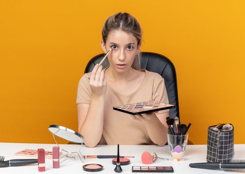 Surprised young beautiful girl sits at table with makeup tools applying eyeshadow with makeup brush isolated on orange background