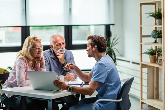 Happy mature couple and their doctor communicating while using laptop during medical appointment