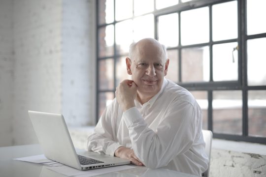 Positive bold aged male freelancer wearing formal shirt sitting at table with laptop and documents and looking at camera with smile during remote work in room with industrial interior