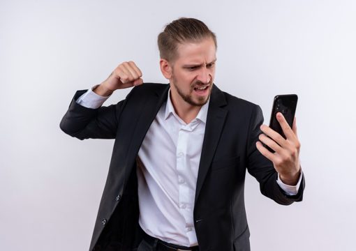 Handsome business man wearing suit holding smartphone looking at screen with angry face with clenched fist standing over white background