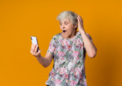 Surprised elderly woman raises hand up looking at phone isolated on orange wall