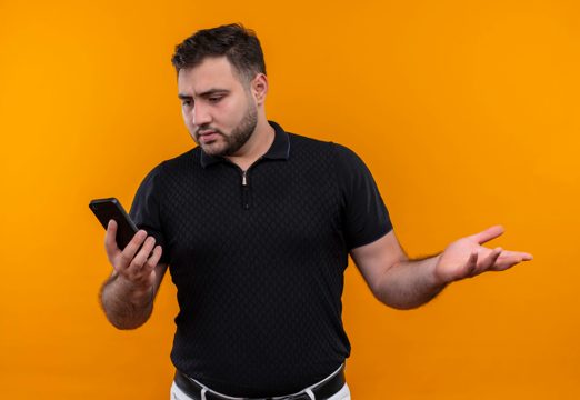 Young bearded man in black shirt holding smartphone   looking confused and very anxious