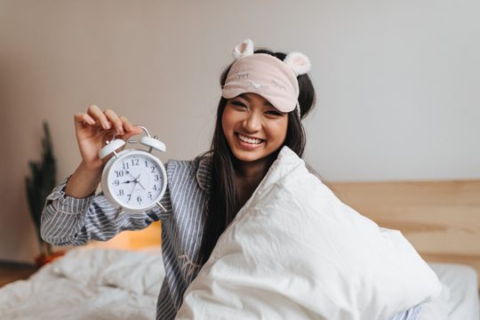 Charming woman in striped pajamas laughs and keeps alarm clock