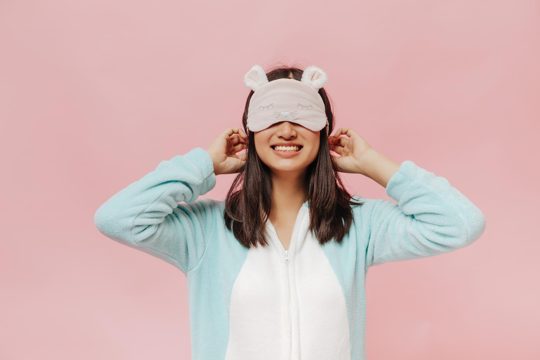 Joyful brunette woman in white and mint soft pajamas puts on cute sleep mask and smiles widely on pink isolated background