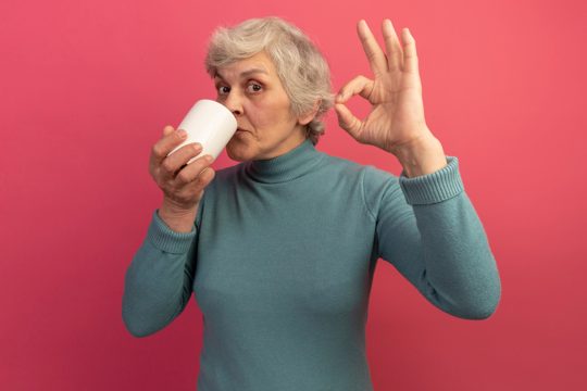 Pleased old woman wearing blue turtleneck sweater drinking cup of tea doing ok sign isolated on pink wall