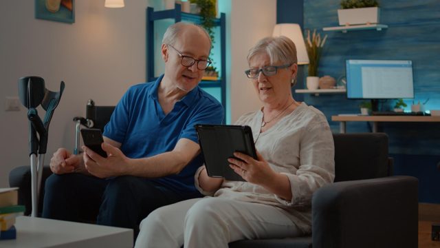 Grandparents using smartphone and digital tablet at home, enjoying technology in free time. man with disability looking at mobile phone screen and starting conversation with wife.