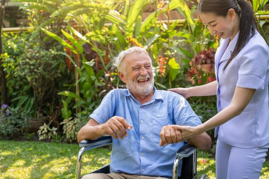 Happy nurse holding laughing elderly man hand on wheelchair in garden at nursing home